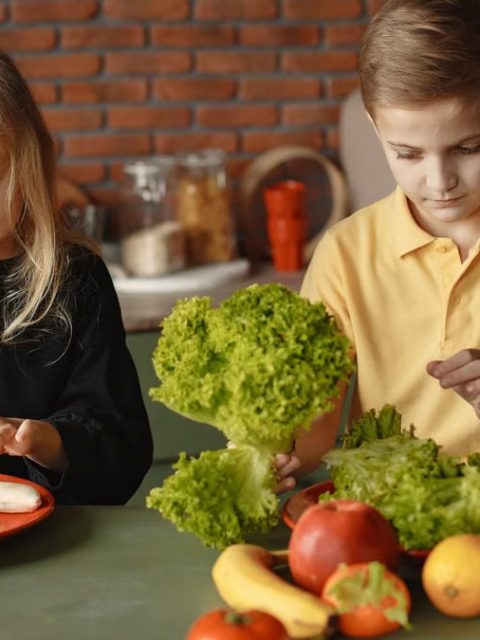 Focused little girl and boy sitting at desk with assorted vegetables and fruits while cooking in loft kitchen with brick wall
